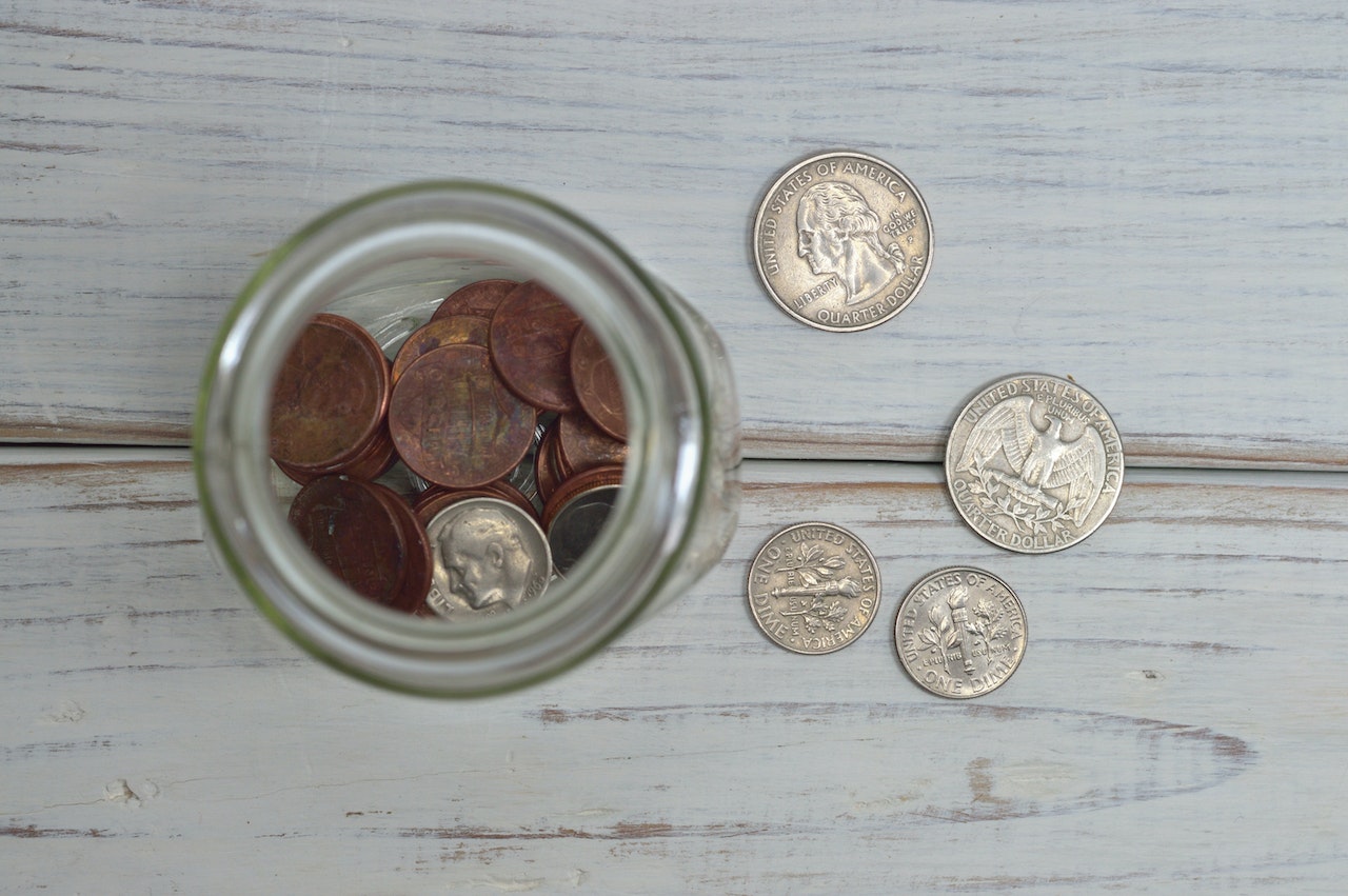 Na zdjęciu widać słoik z monetami o różnych nominałach. Monety są ułożone jedna na drugiej, tworząc wrażenie masy pieniędzy zgromadzonych w jednym miejscu. Słoik, w którym znajdują się monety, jest przezroczysty, co pozwala na łatwe zobaczenie zawartości